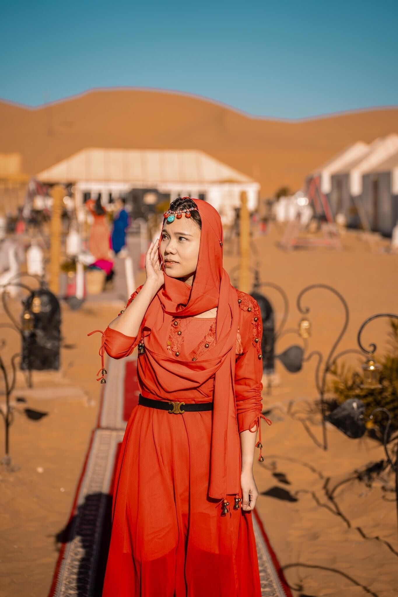 A person in a red outfit standing in a desert setting with tents and a market in the background.