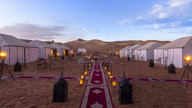 A desert camp with tents arranged in rows, a central walkway with lanterns, and a clear sky.