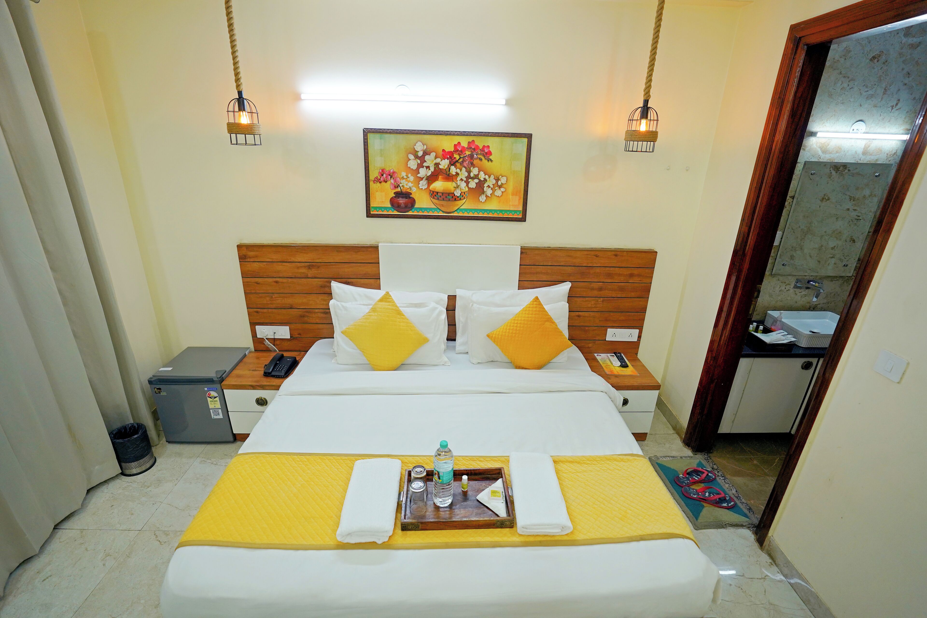 A hotel room with a bed, bedside tables, a nightstand, a trash bin, a wall-mounted painting, and a door leading to a bathroom.