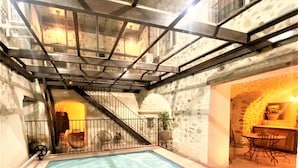 An indoor swimming pool with a stone wall backdrop, a staircase leading to an upper level, and a seating area with tables and chairs. - Gîte 4* for 6p, 3ch, 100m2, HEATED SWIMMING POOL, PATIO (Vallon pont d'arc)