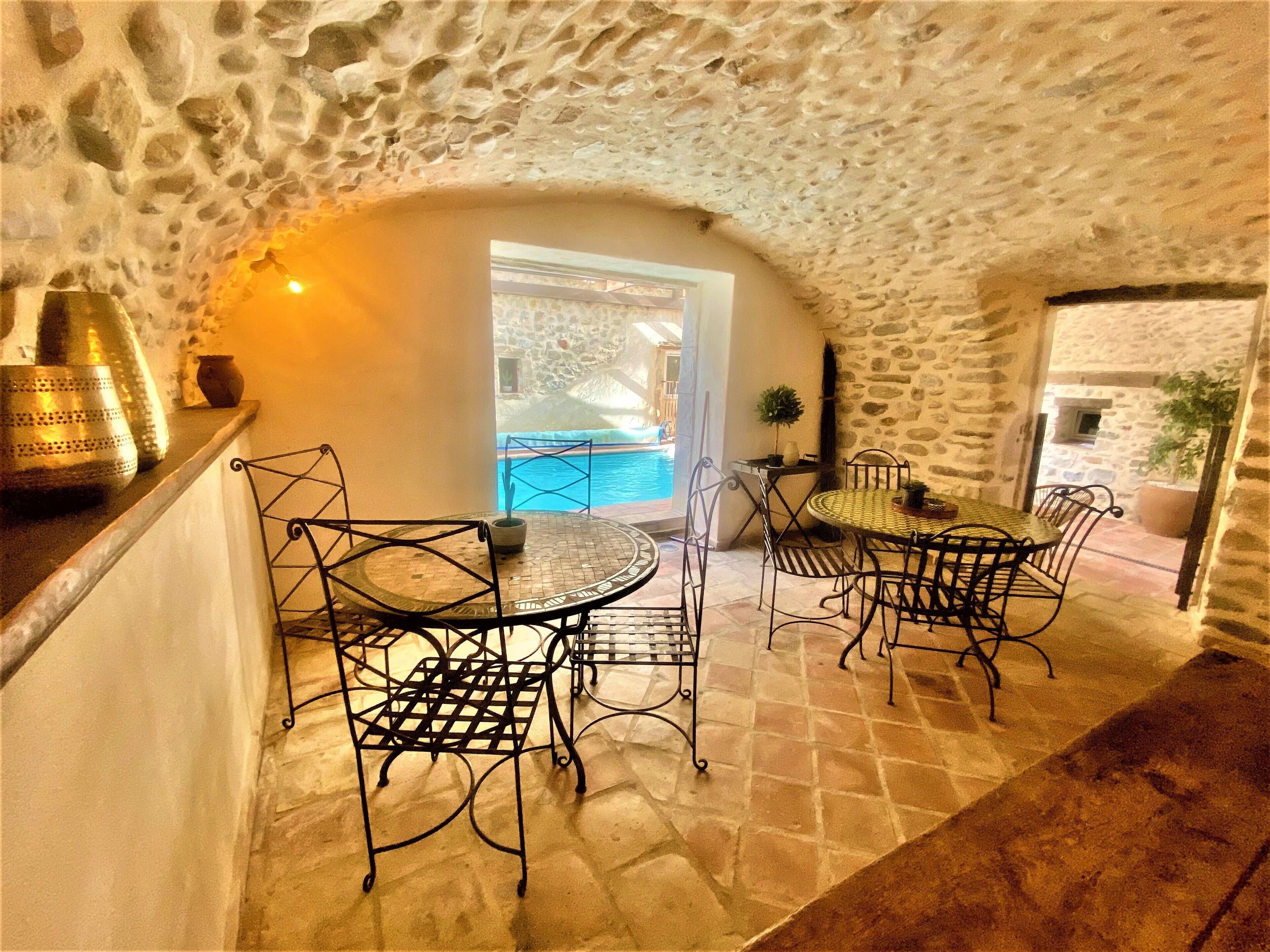 Un espace repas aux murs de pierre, avec vue sur la piscine, des chaises en métal et une table recouverte d’une nappe à motifs.