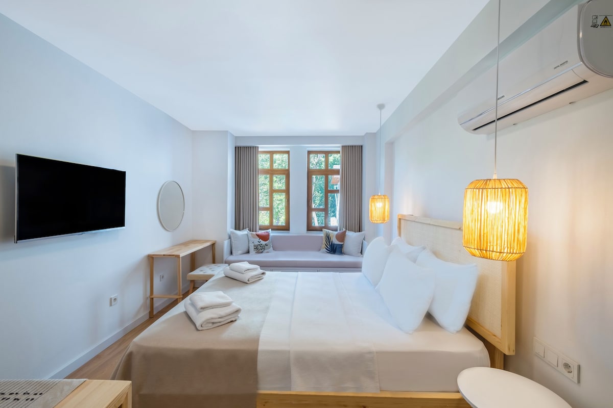A hotel room with a bed, a TV, a window with curtains, a bedside table, and a lamp.