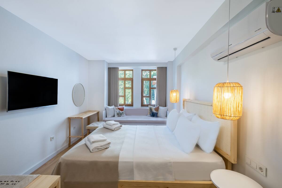 A modern hotel room with a large bed, a TV mounted on the wall, a window seat with curtains, and a hanging lamp.