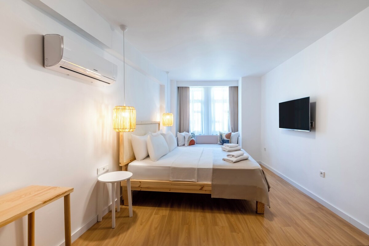 A minimalist hotel room with a bed, bedside lamps, a TV, and a window with curtains.