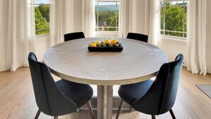 Una mesa de comedor redonda con acabado en madera clara, rodeada por cuatro sillas negras con patas de madera. Un candelabro con adornos similares a astas cuelga del techo. La habitación tiene ventanas amplias con cortinas blancas, que ofrecen vistas a la vegetación y un cielo despejado.