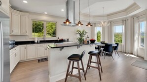 Una cocina moderna con muebles blancos, una isla central con taburetes de bar, iluminación colgante y vistas a zonas verdes.