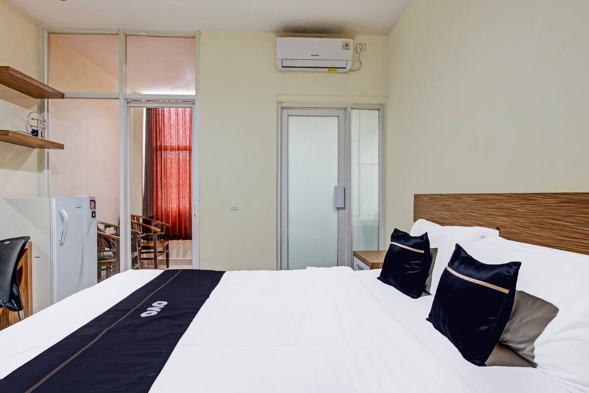 A hotel room with a bed, a desk, a chair, a refrigerator, and a sliding door to a balcony.