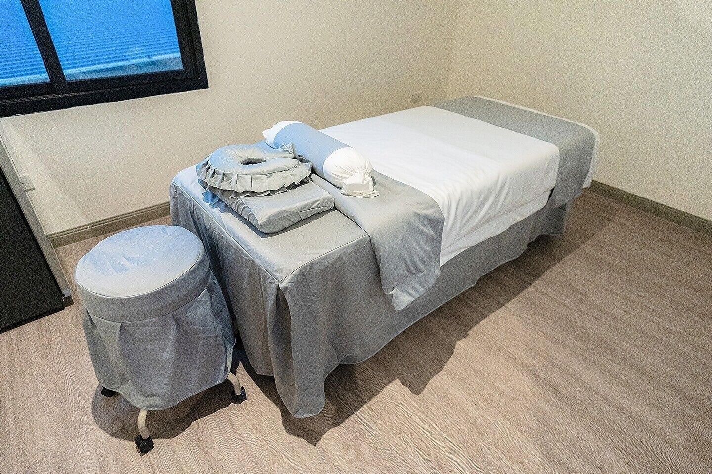 Ein Einzelbett mit weißer Bettdecke, ein kleiner runder Hocker und ein Kopfkissen auf einem Tisch.