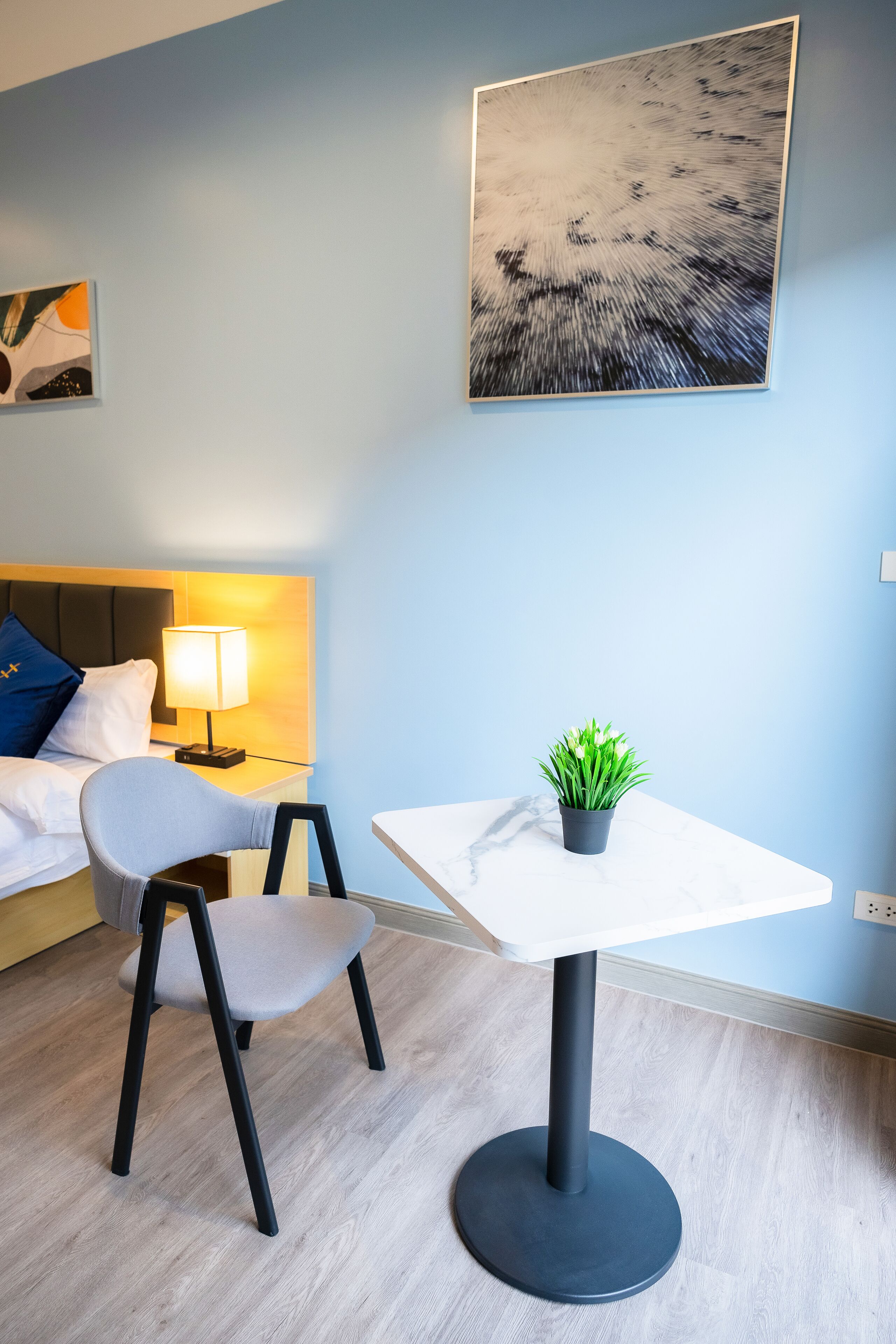 A modern hotel room with a bed, bedside table with a lamp, a small table with a plant, and a painting on the wall.