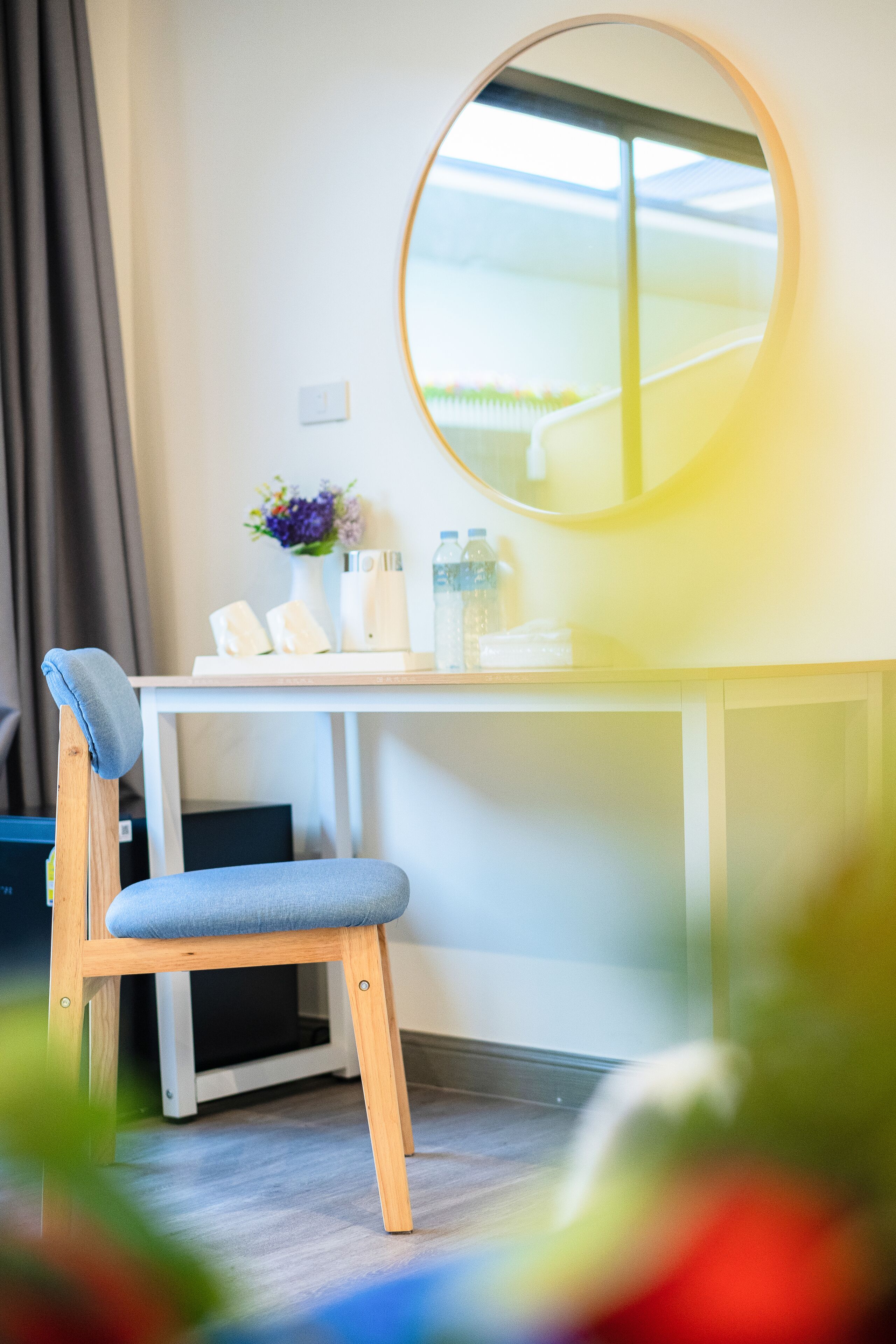 Une chambre moderne avec une chaise en bois, un bureau, un miroir rond et une fenêtre donnant sur un paysage verdoyant.