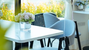 A modern dining area with a white table and two grey chairs, a vase with flowers, and a view of greenery outside. - Fresh Resort Pattaya (Pattaya)