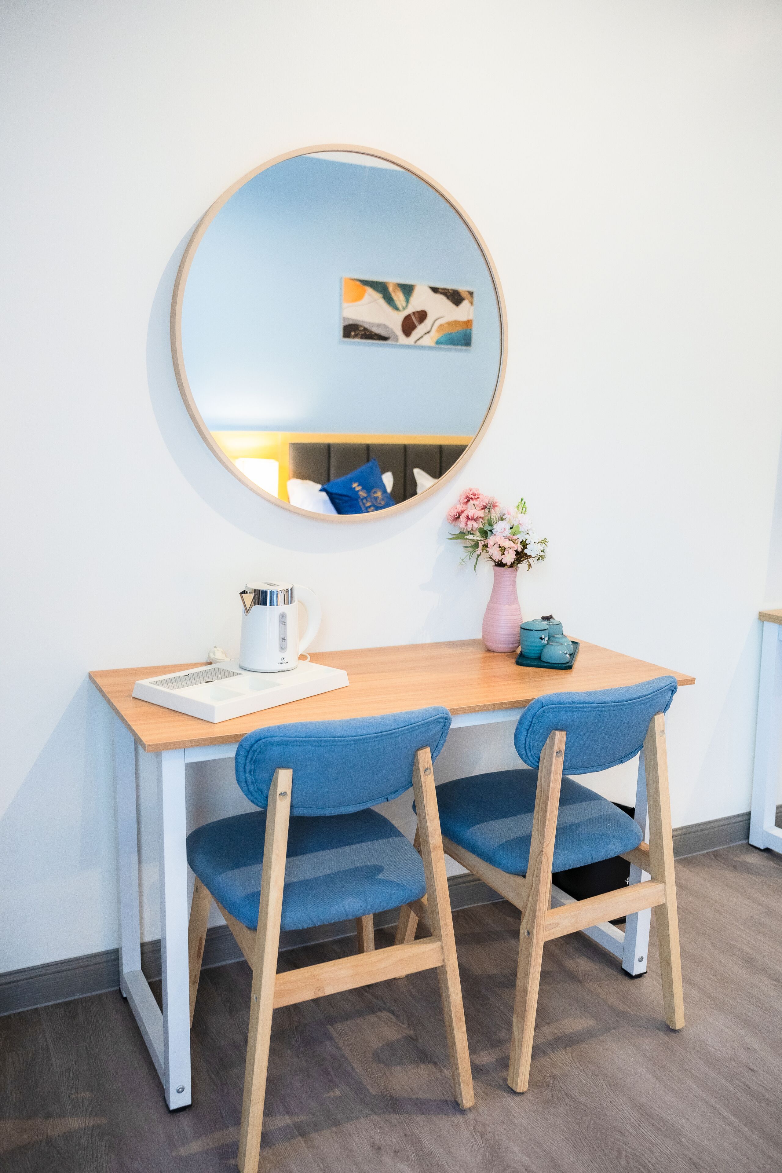 A small, modern room with a wooden table, two blue chairs, a white kettle, a pink vase with flowers, and a round mirror on the wall.
