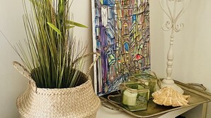 A room with a woven basket containing green plants, a colorful abstract painting, a white metal stand, and a glass jar with a candle.