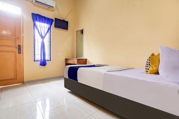 A hotel room with a bed, a window with blue curtains, a bedside table, a mirror, and a wall-mounted TV.
