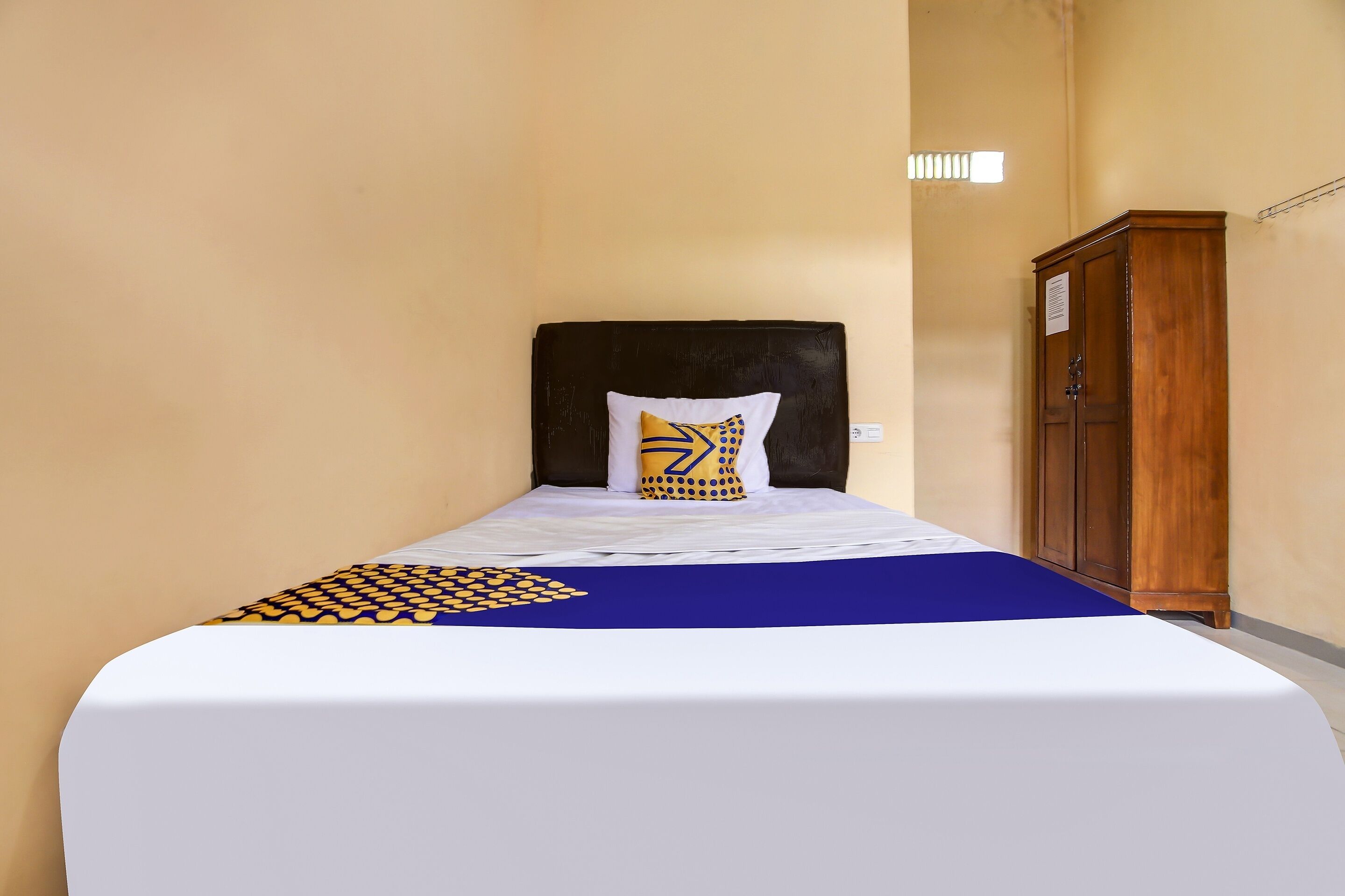 Una cama bien hecha con una colcha blanca y azul, una almohada estampada y un armario de madera frente a una pared lisa.