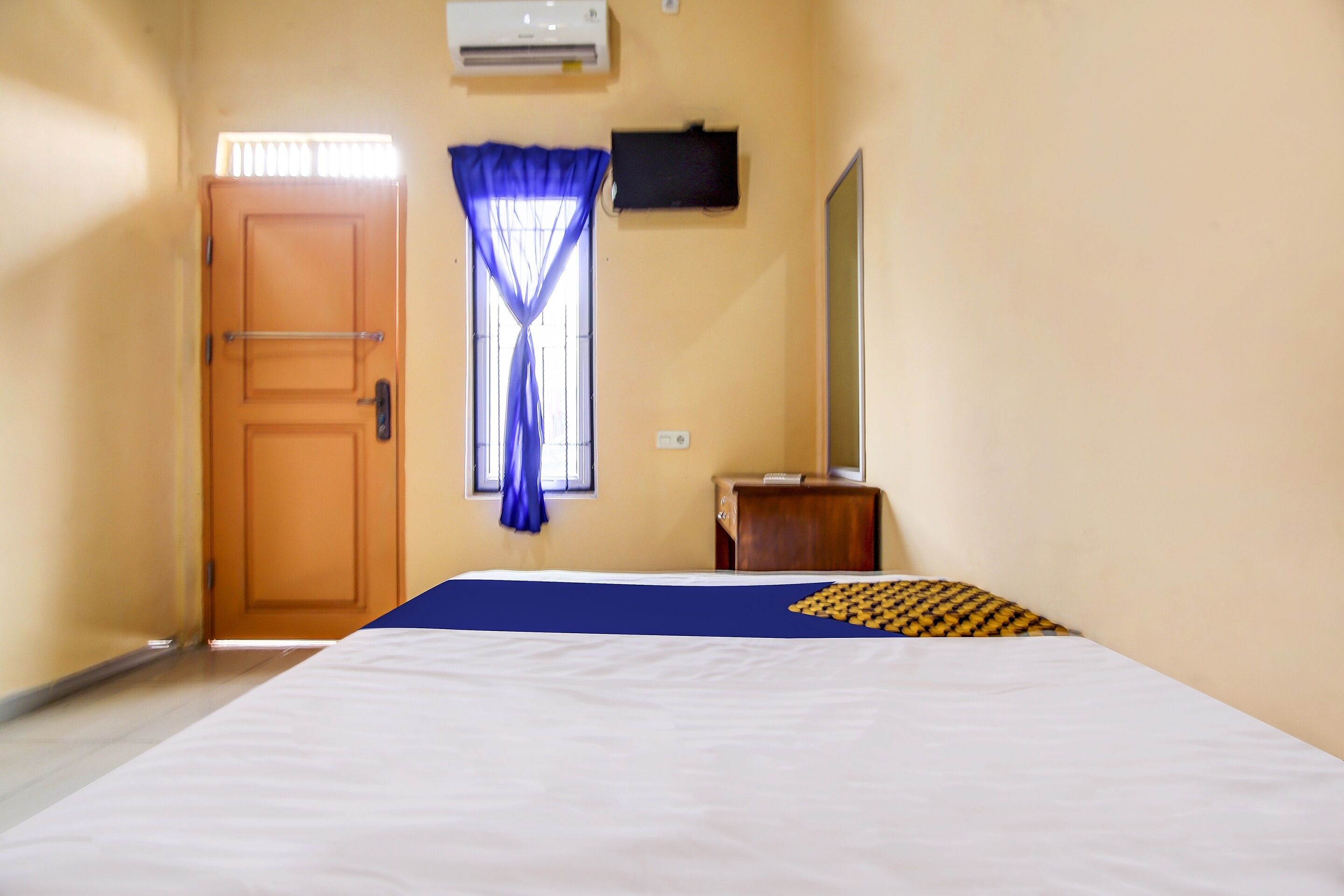 A hotel room with a bed, a wooden door, a window with blue curtains, a wall-mounted TV, and a small wooden cabinet.