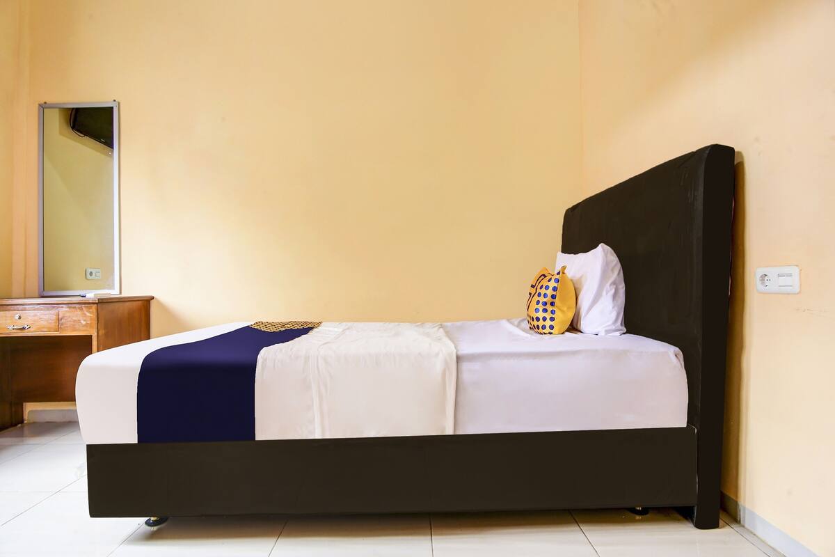 A neatly made bed with a dark headboard, a wooden nightstand with a mirror, and a plain wall.