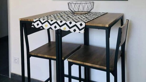 A dining area with a table and chairs, a geometric patterned tablecloth, a black metal-framed mirror, and a hanging light fixture. - F3 Lumineux Tout Équipé sur la Route des Plages (Perpignan)
