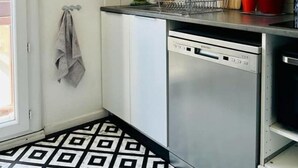 A kitchen with white cabinets, a dishwasher, a sink, and a patterned floor. There are signs for coffee and hot dogs on the wall. - F3 Lumineux Tout Équipé sur la Route des Plages (Perpignan)