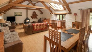 A spacious loft with high ceilings, wooden beams, a fireplace, a dining area, and a cozy seating area. - Cowslip Barn (Totnes)