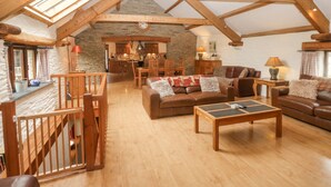 A spacious loft with a high ceiling, exposed wooden beams, a stone wall, and a cozy seating area with a fireplace. - Cowslip Barn (Totnes)