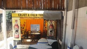 A covered outdoor dining area with a long table and chairs, a sign advertising a cold beverage, and a rustic wooden ceiling. - Hotel Bison (Nakuru)