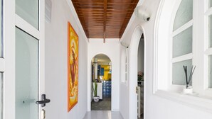 A corridor with a tiled floor, white walls, and a wooden ceiling. There are two doors on the left, one with a glass panel and a handle, and another with a metal handle. A large painting is on the wall to the left of the corridor. - Puerto del Carmen Vacation Apartment (Puerto del Carmen)