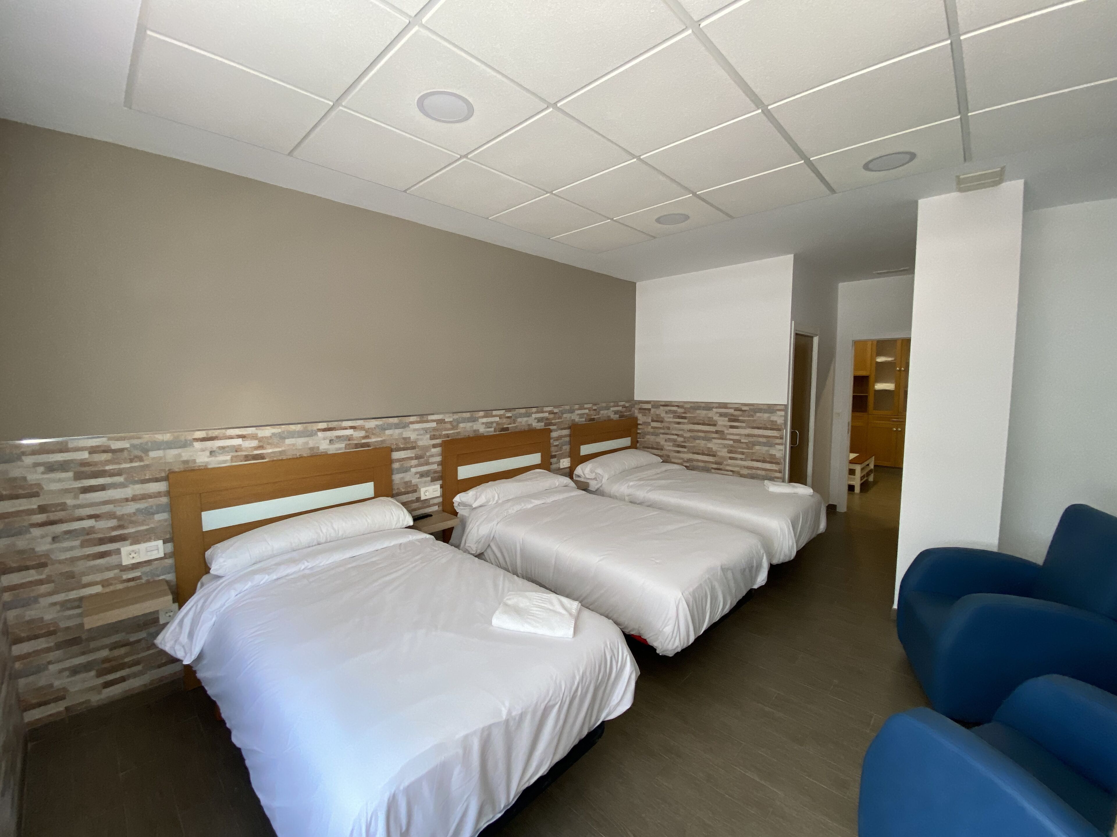 A hotel room with three beds, a blue chair, and a hallway leading to a kitchen area.