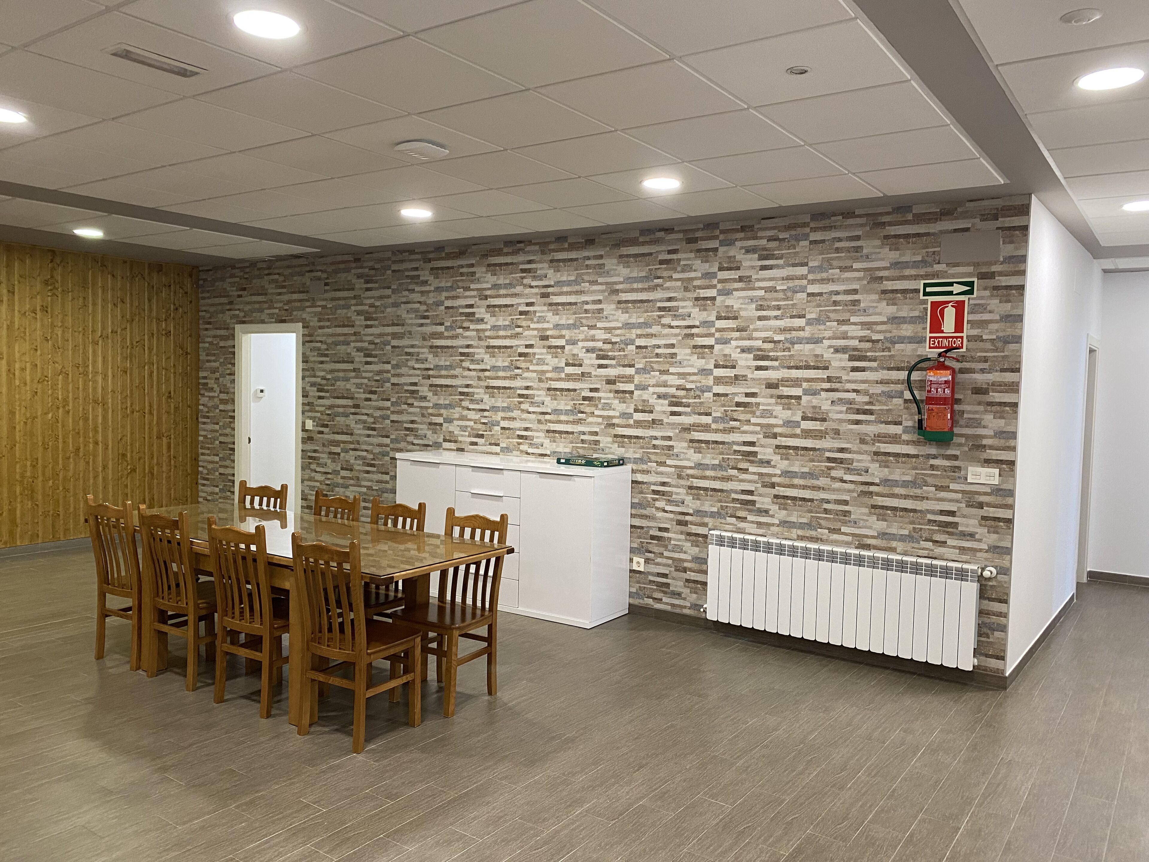 Una sala de conferències amb taula i cadires de fusta, un armari blanc, una paret amb relleu de pedra, un extintor i un radiador.