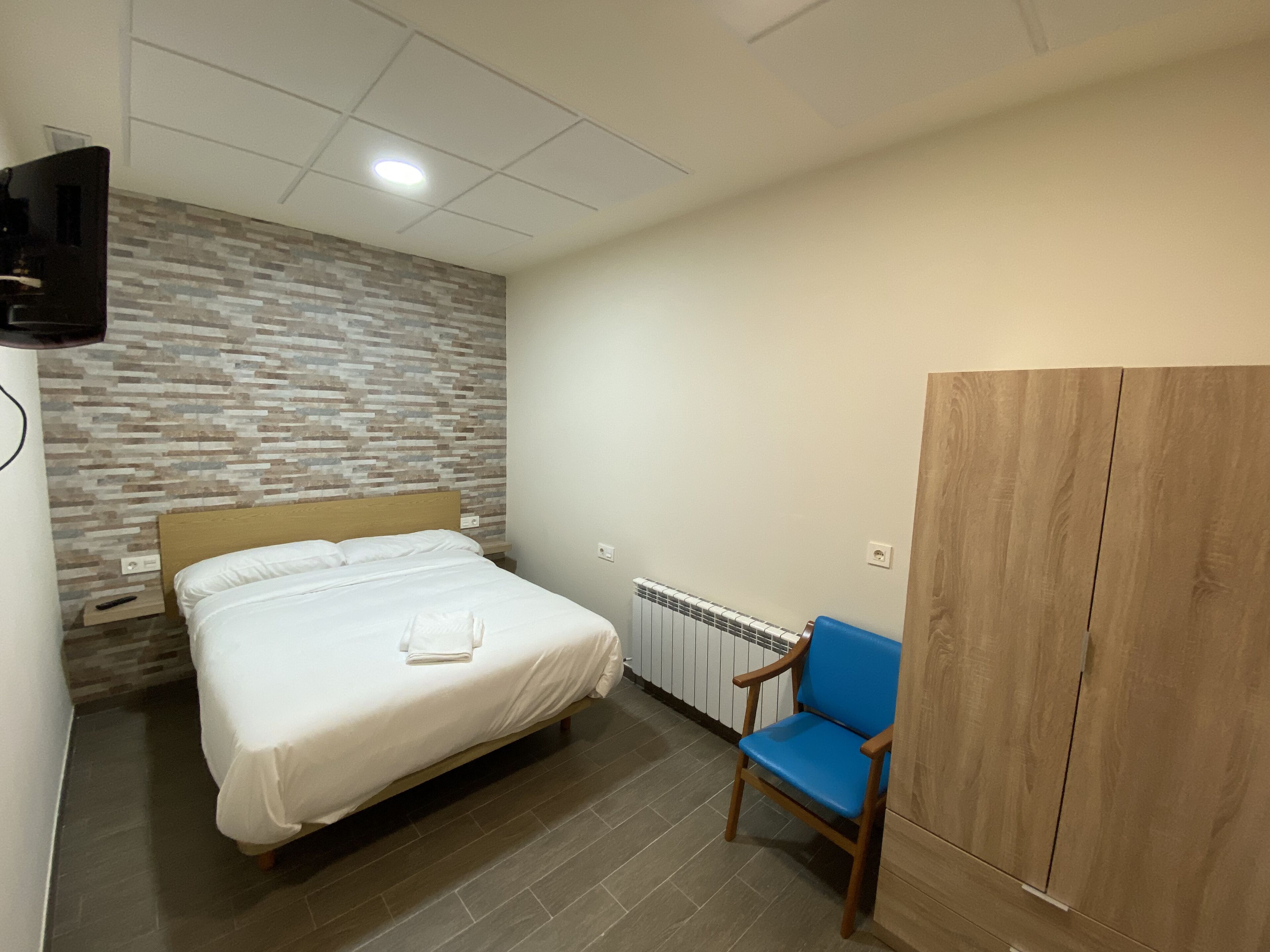 Un dormitorio con una cama, un sillón azul, un armario de madera, un radiador y un televisor fijado a la pared.