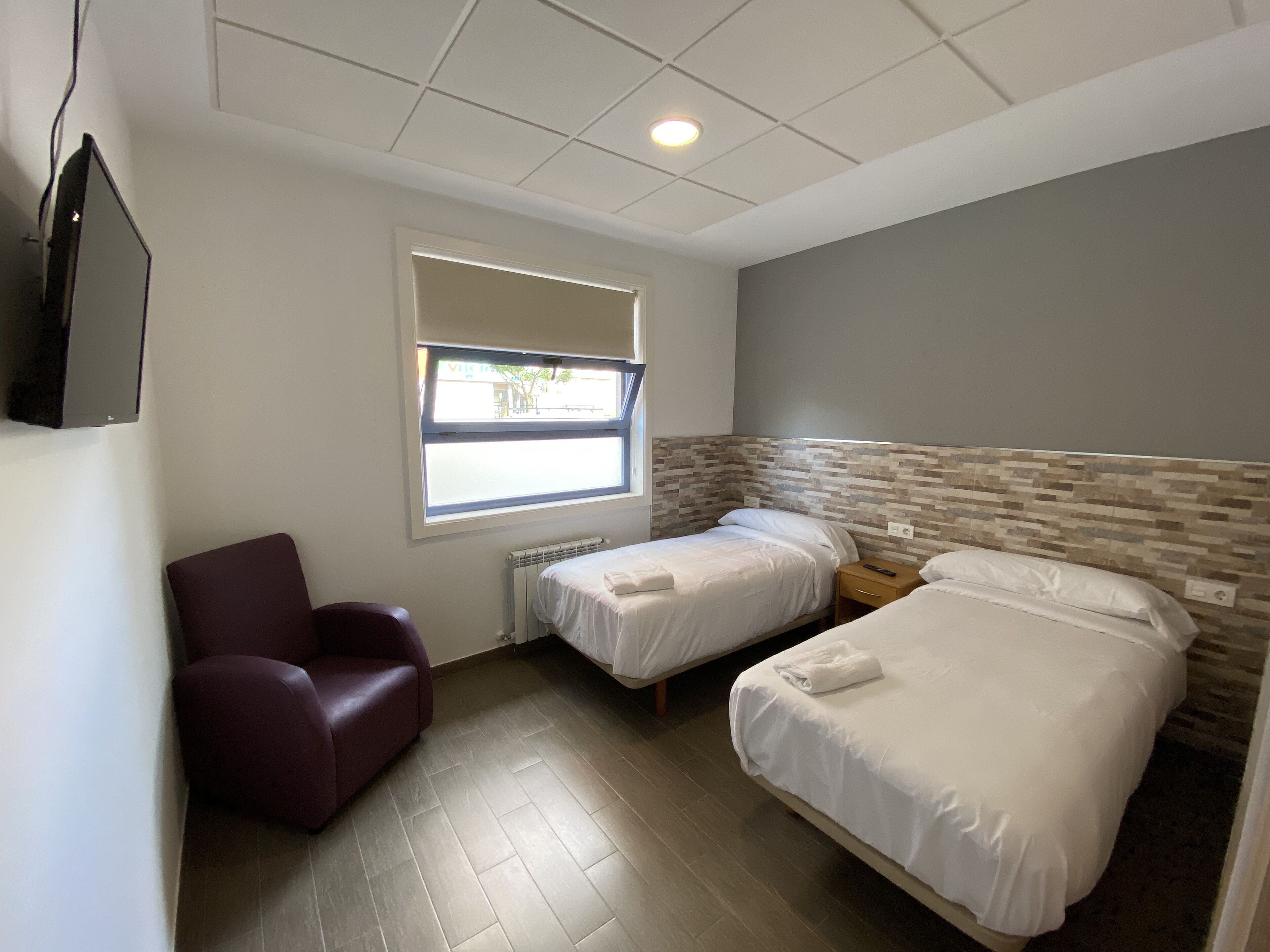 Una camera d'albergo con due letti, una sedia, una finestra e un pavimento piastrellato.