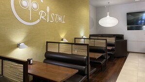 Interior de un restaurante con diseño moderno, que incluye un logo grande en la pared, mesas de madera y asientos tipo banca acolchados en negro, además de una lámpara colgante.