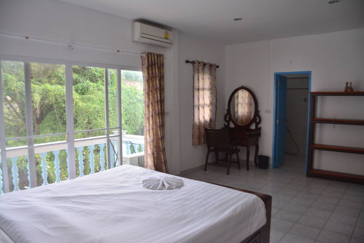 A hotel room with a bed, a desk, a chair, a mirror, and a view of greenery outside.