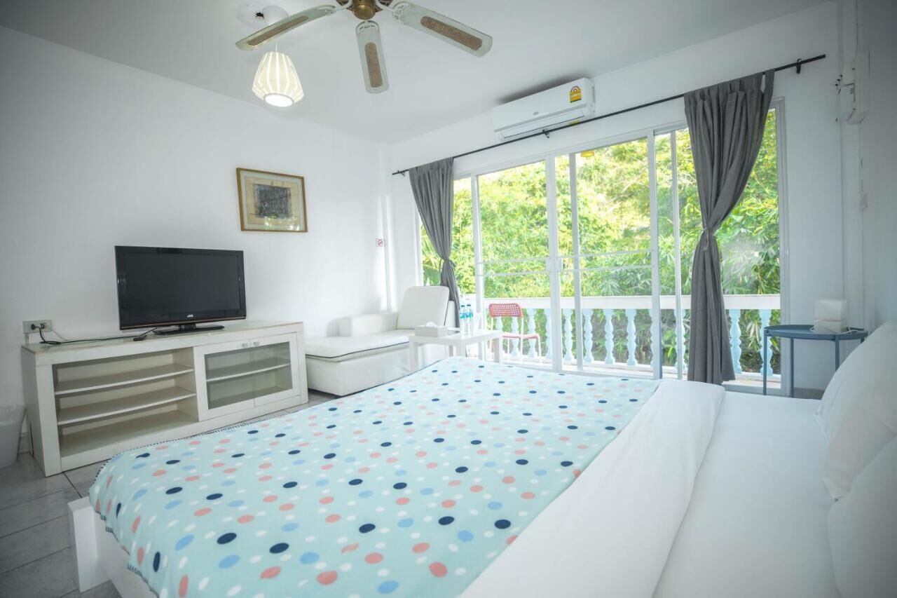 A bedroom with a bed, a TV on a white cabinet, a balcony with a chair, and a large window with curtains.