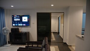 Un salon moderne avec un téléviseur à écran plat fixé au mur, un banc en bois, un ventilateur sur pied et un canapé aux couleurs sombres.