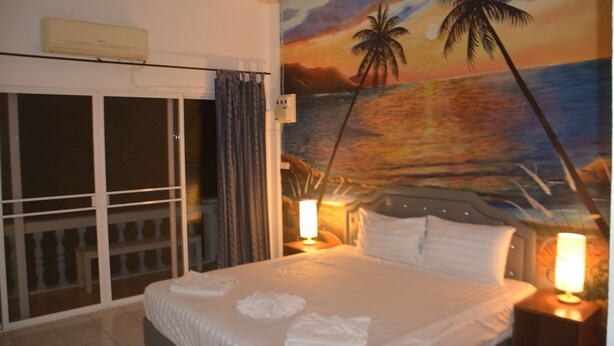 Une chambre d’hôtel avec un lit, un tableau représentant un coucher de soleil, une fenêtre avec des rideaux et une table de chevet avec une lampe.