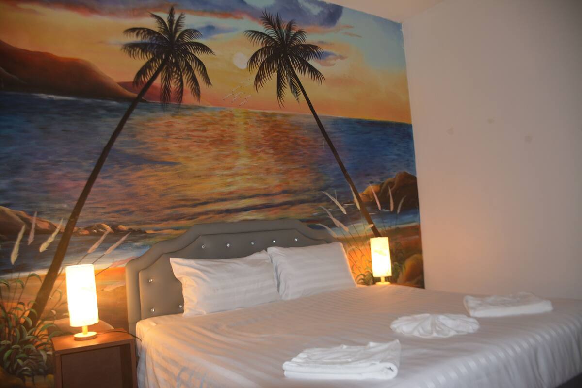 A hotel room with a bed, two bedside lamps, and a mural of a beach scene.