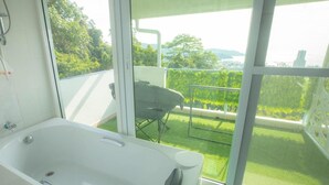 Une salle de bain moderne dotée d’une grande baignoire, d’une douche et d’un balcon offrant une vue sur un paysage verdoyant et des bâtiments éloignés.