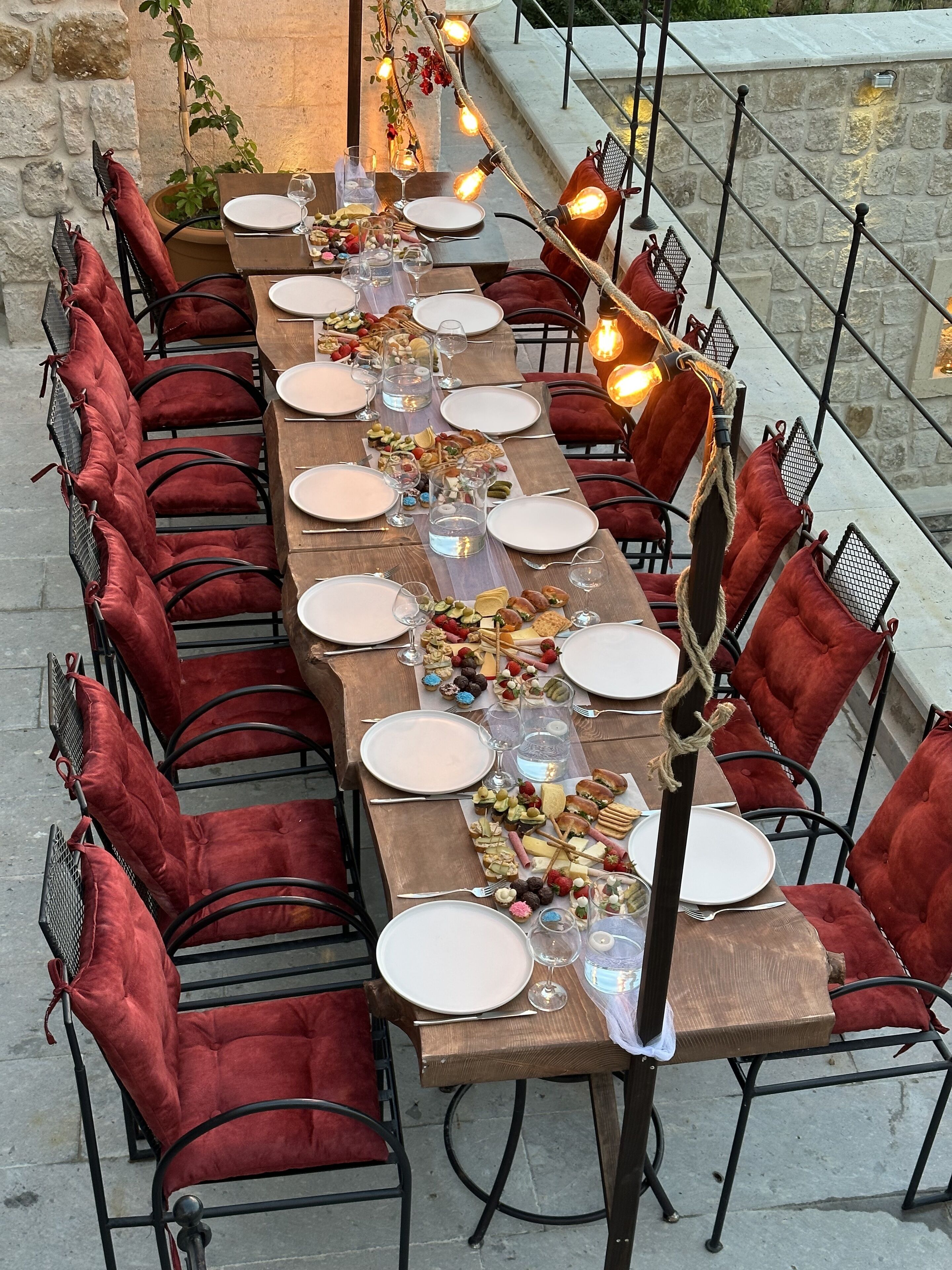 Una larga mesa de comedor dispuesta con platos, vasos y diversos alimentos, rodeada de sillas con cojines rojos.