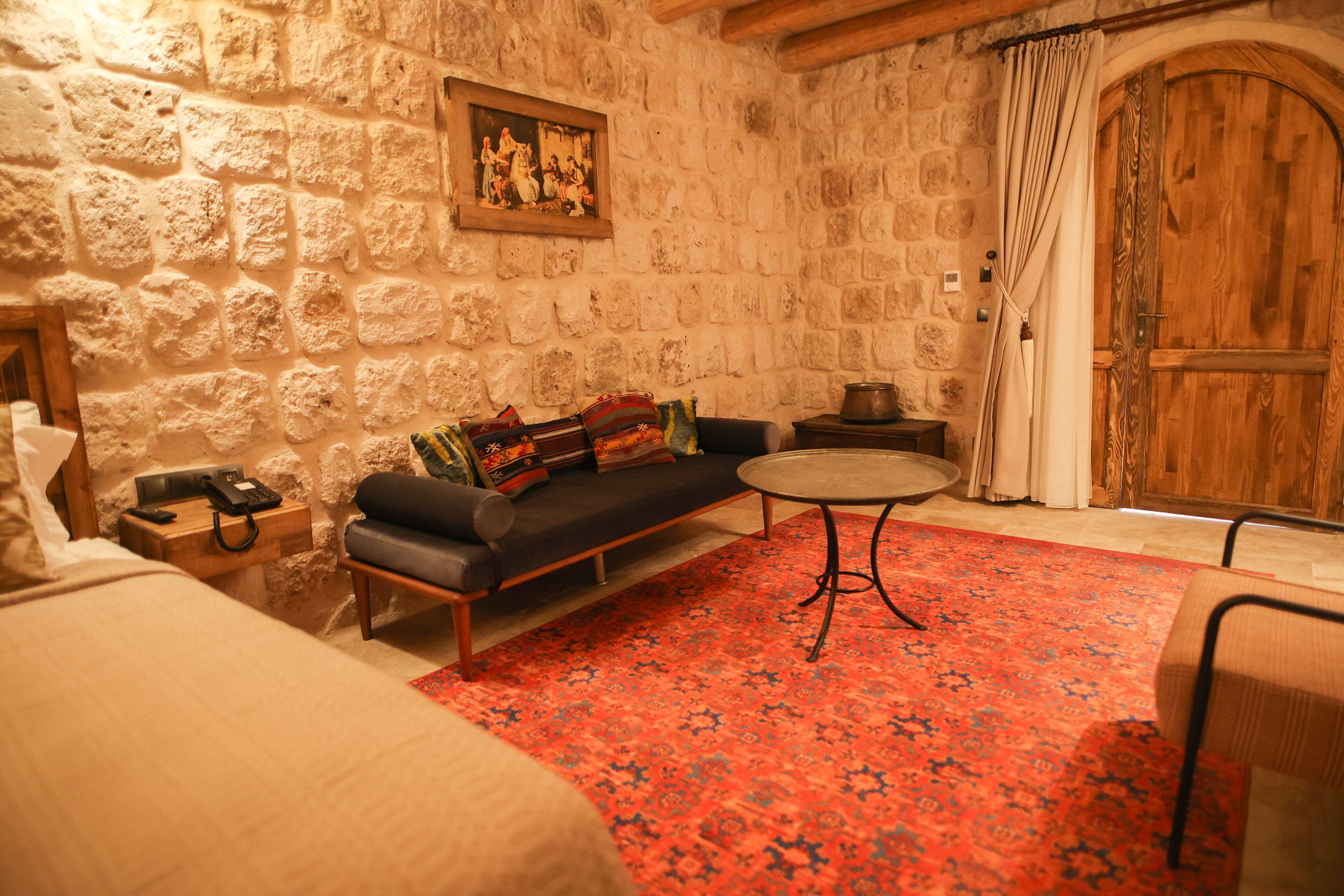 Una habitación acogedora con pared de piedra, puerta de madera, sofá con cojines de colores, una mesita redonda y alfombra estampada.