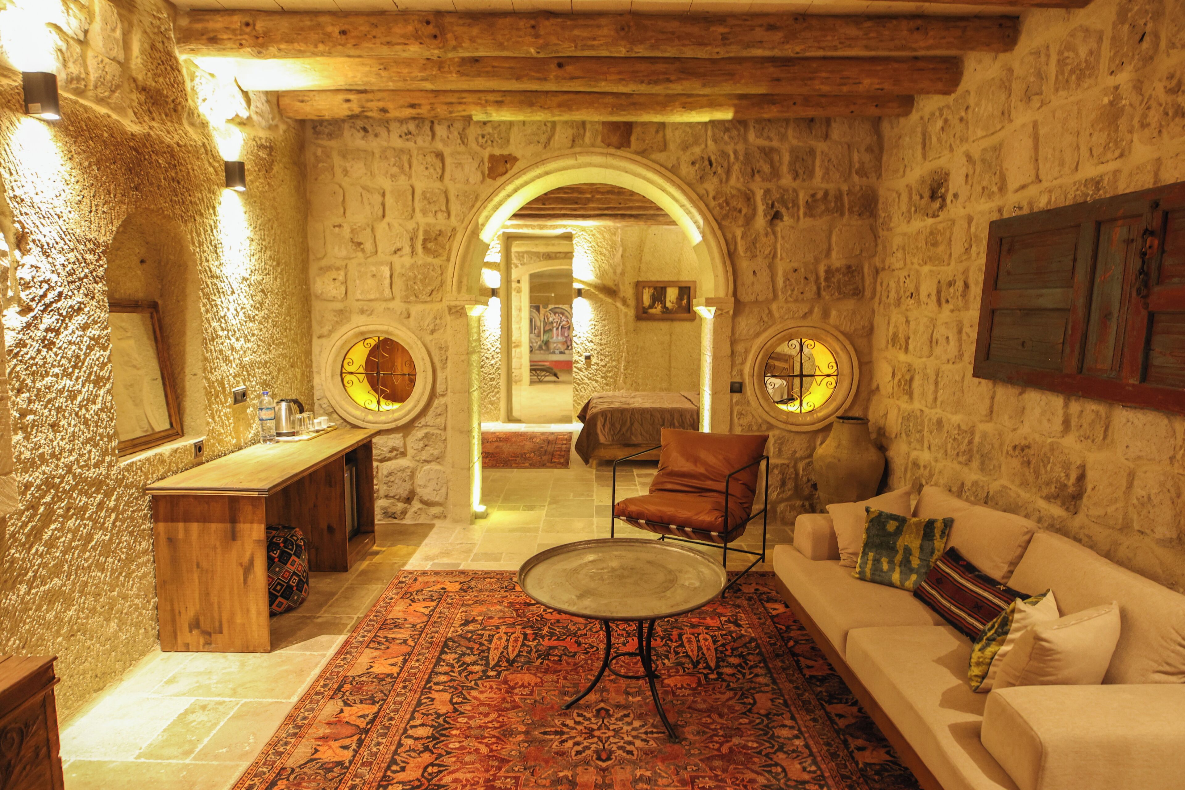 Una habitación de paredes de piedra con un banco de madera, una mesa redonda, un sofá y una alfombra estampada.