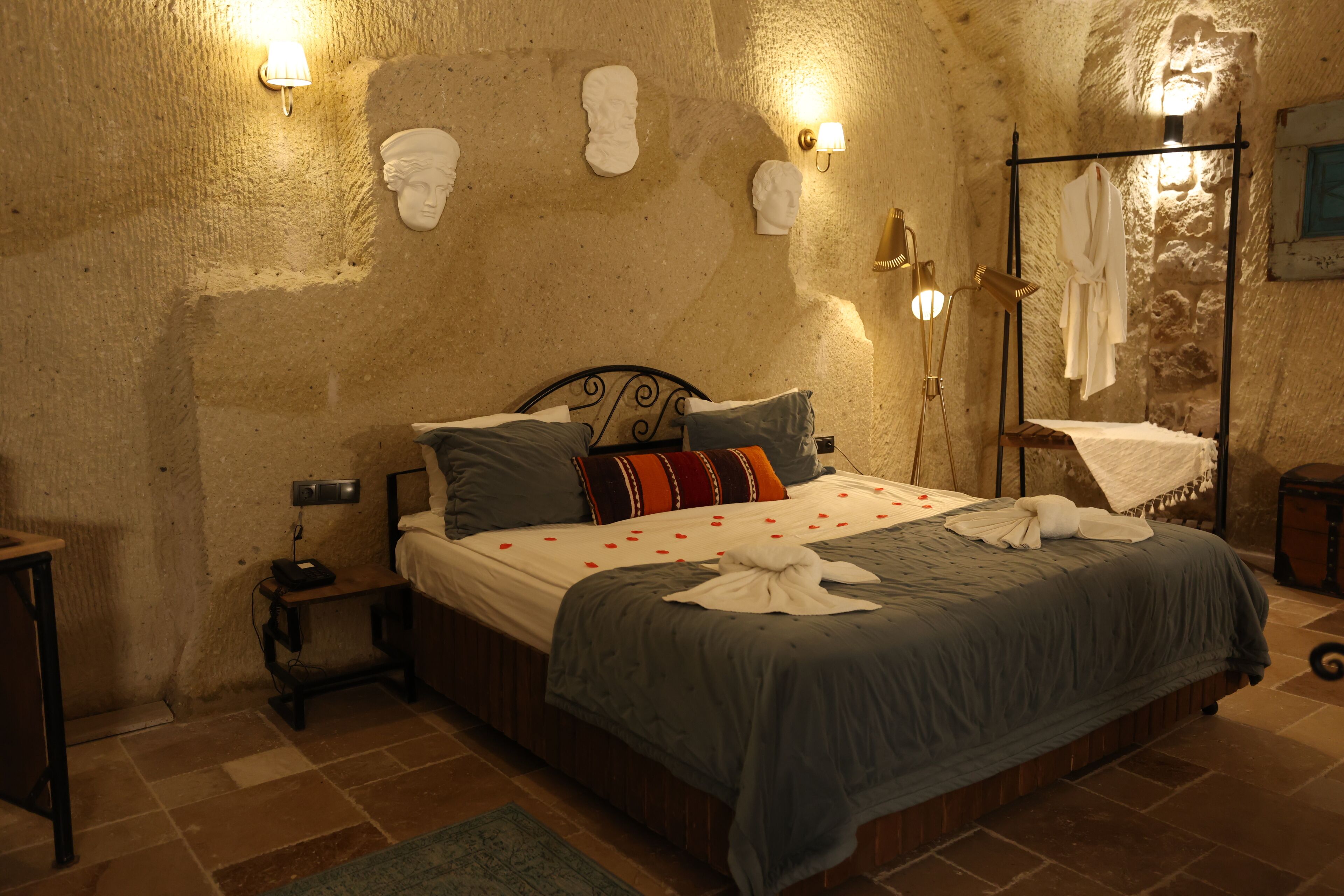 A bedroom with a bed, headboard, bedside tables, wall-mounted lights, and a stone wall.