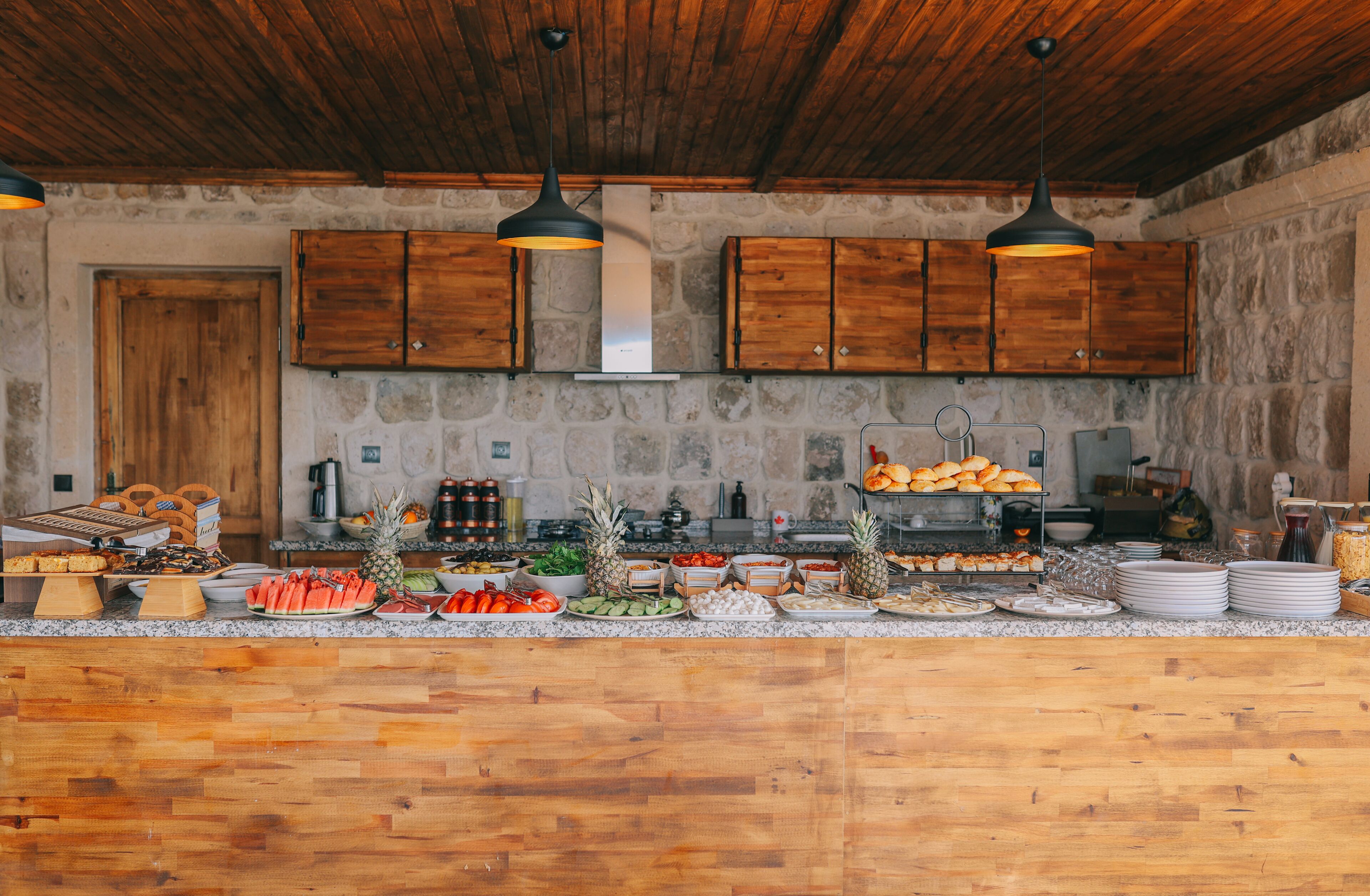 A rustic kitchen with wooden cabinets, a stone wall, and a variety of food items displayed on the counter.