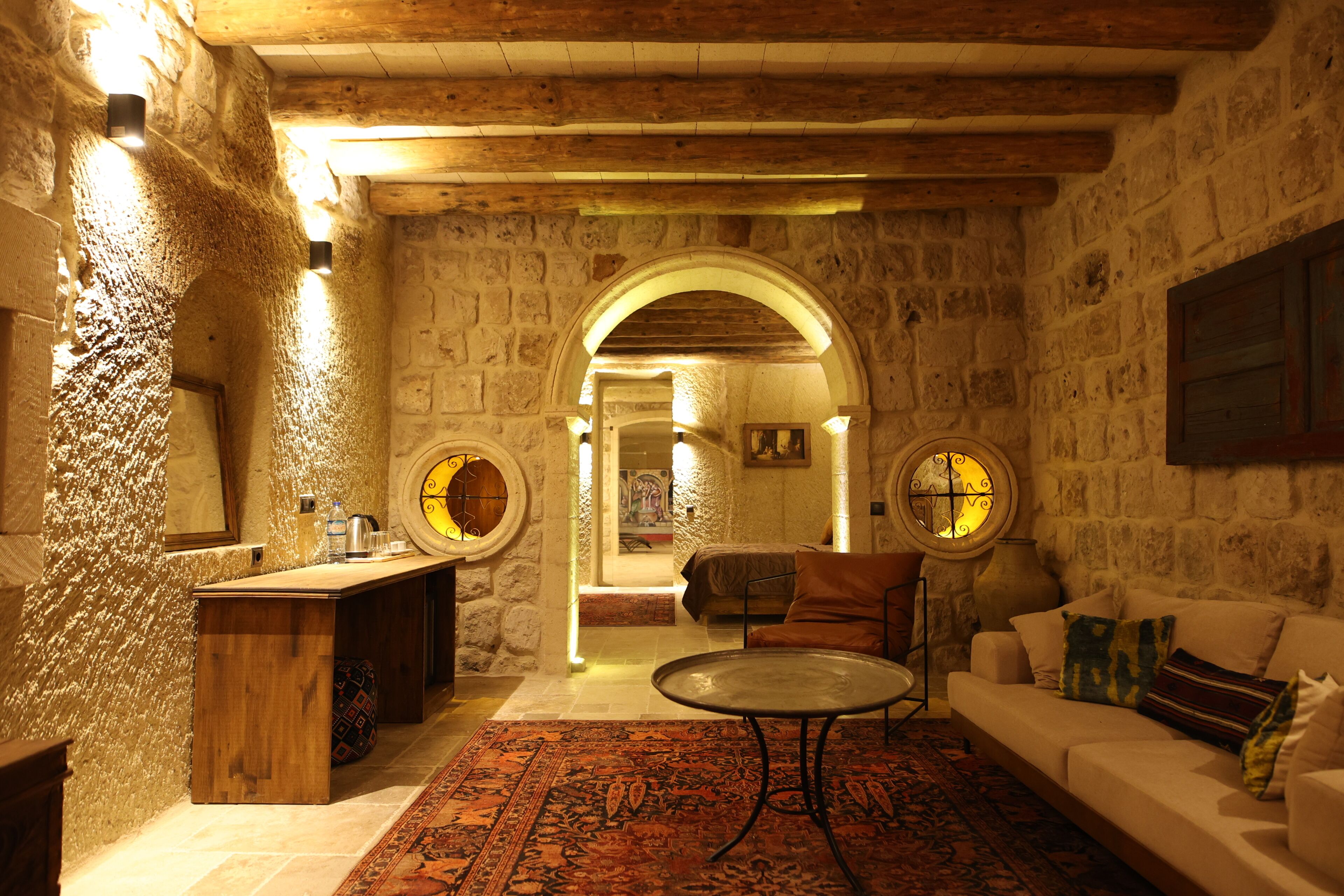 A stone-walled room with a wooden ceiling, a round table, a sofa, and a small bar area.