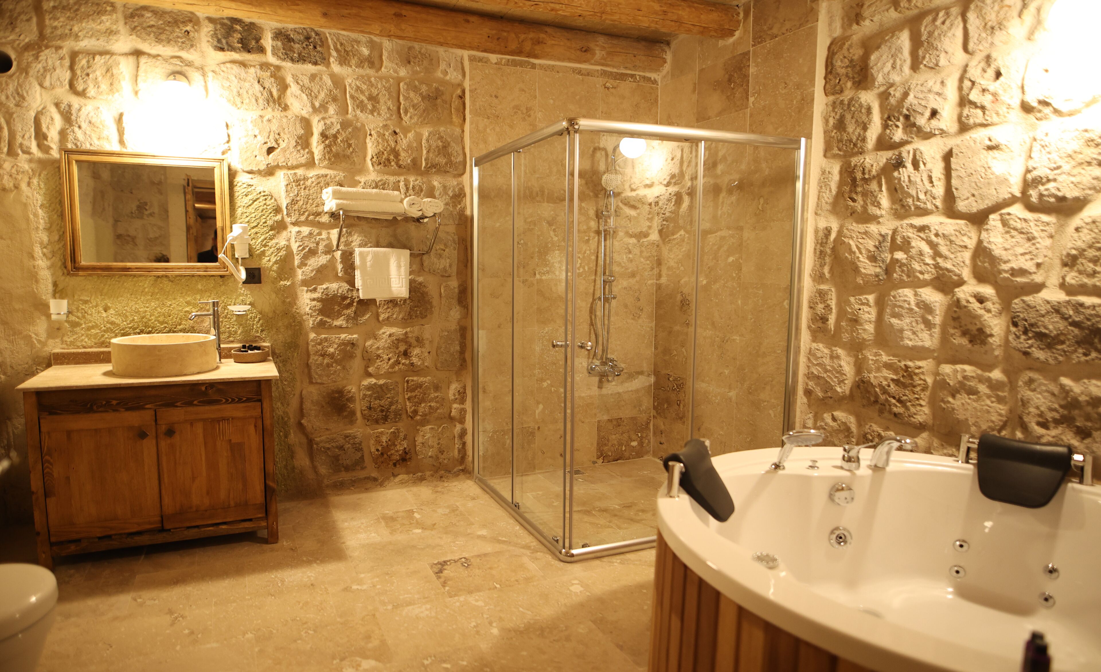 A bathroom with a stone wall, a wooden vanity, a glass-enclosed shower, and a freestanding bathtub.
