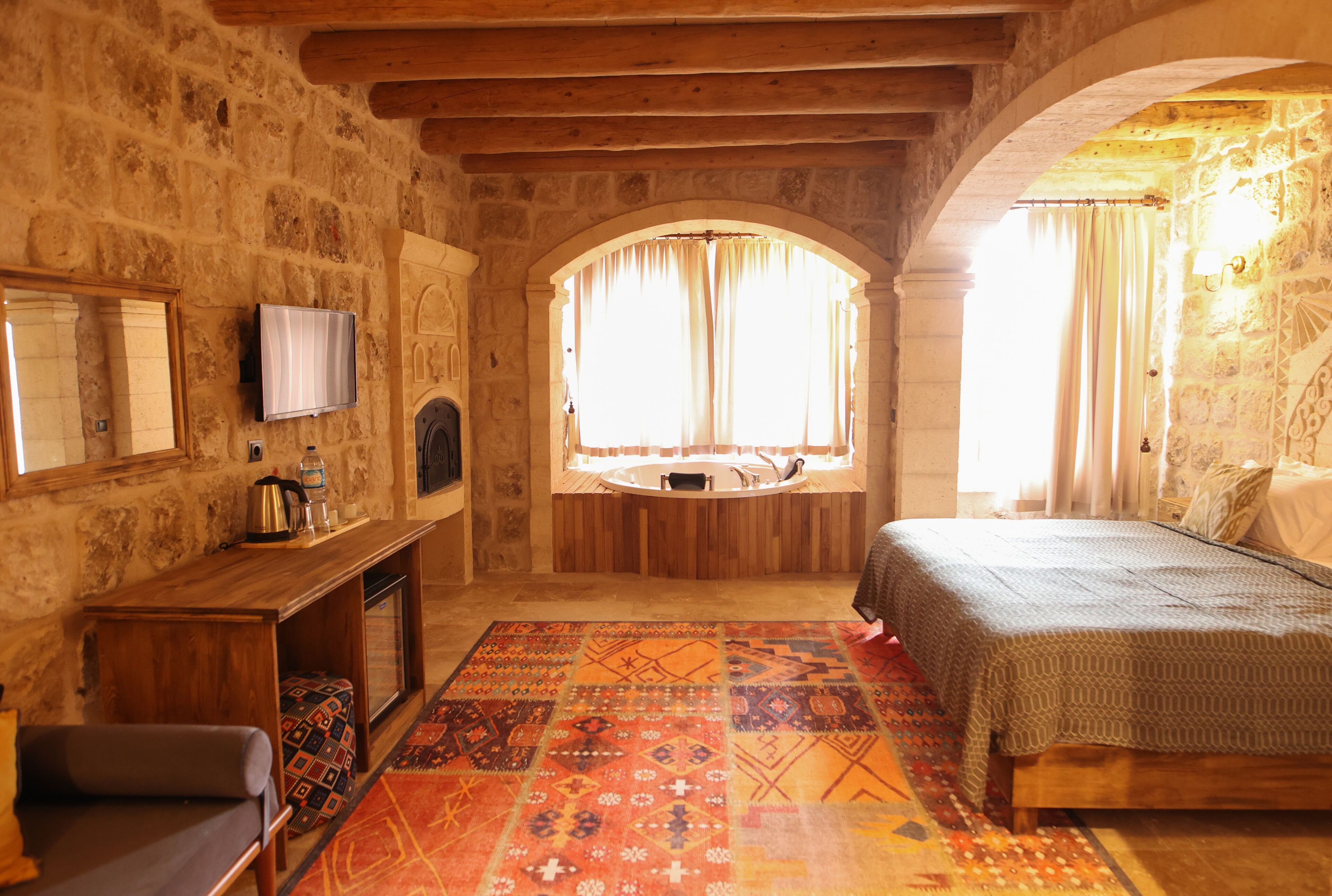 Un dormitorio con pared de piedra, vigas de techo de madera, un ventanal con cortinas, una cama, un televisor y un banco de madera.