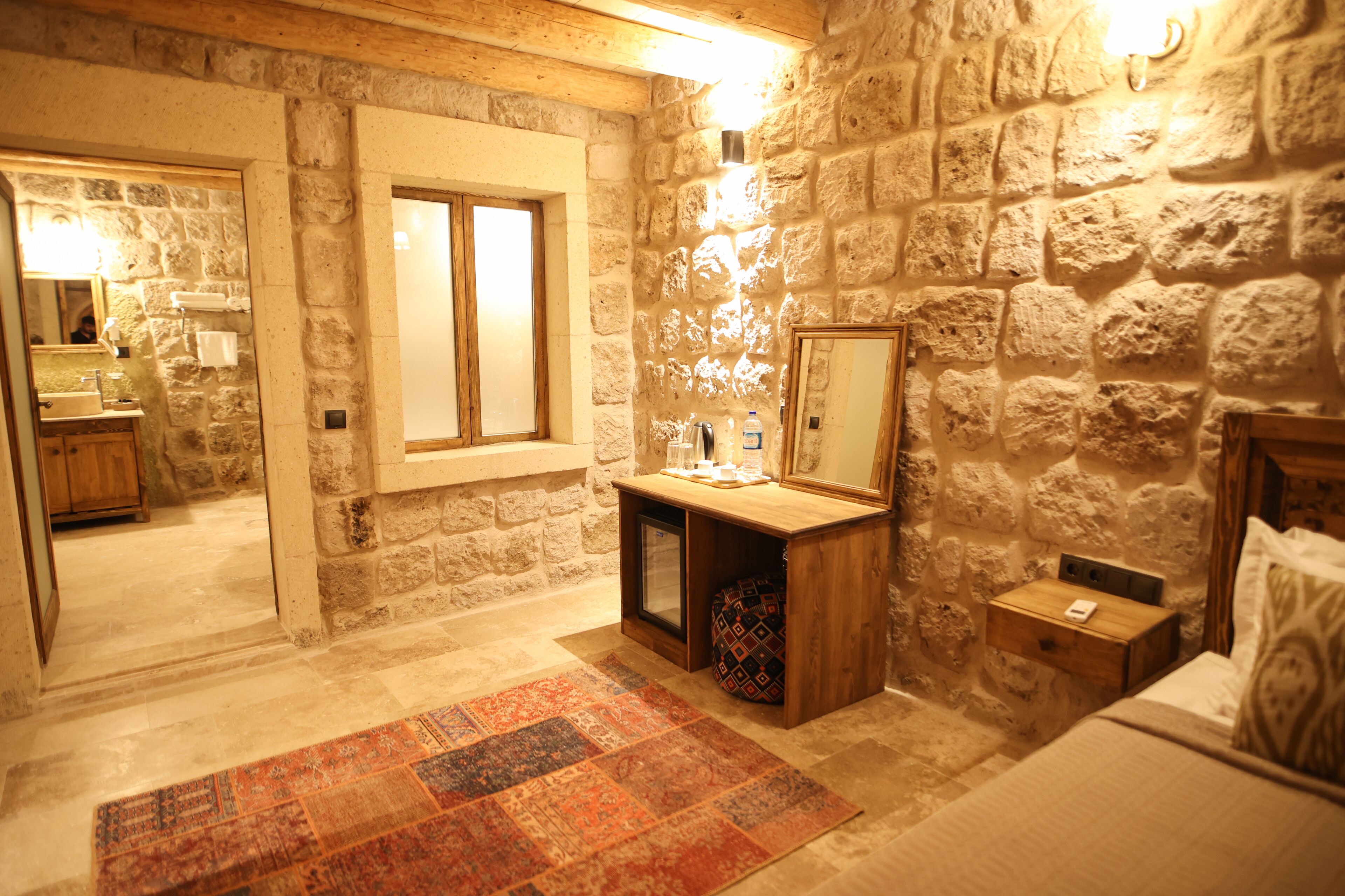A stone-walled room with a bed, a nightstand, a wooden dresser, a mirror, and a window.