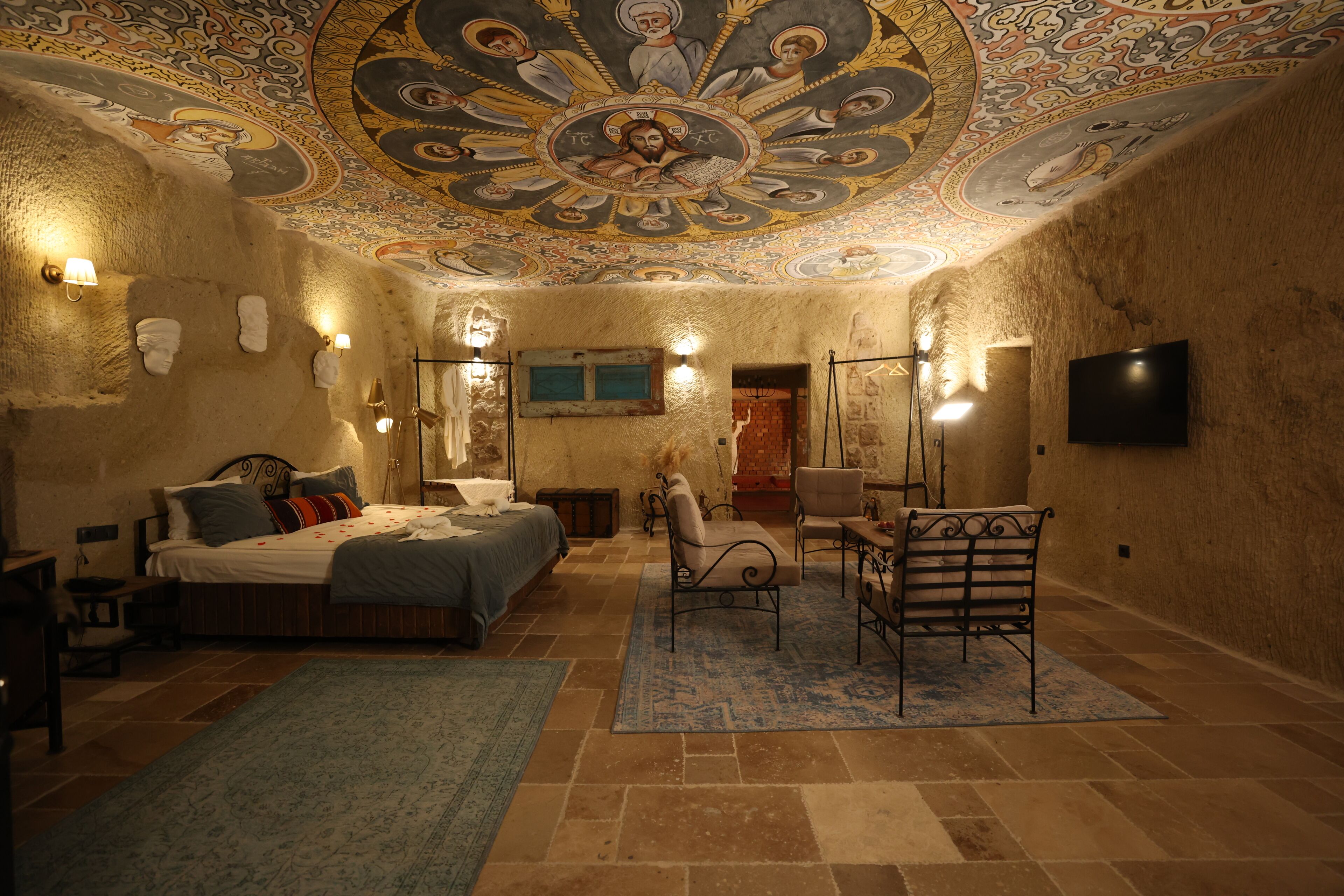 A room with a bed, a chair, a desk, a television, and a ceiling with intricate artwork.