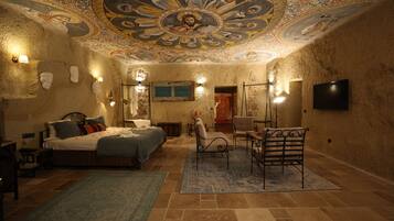 A room with a bed, a chair, a desk, a television, and a ceiling with intricate artwork.