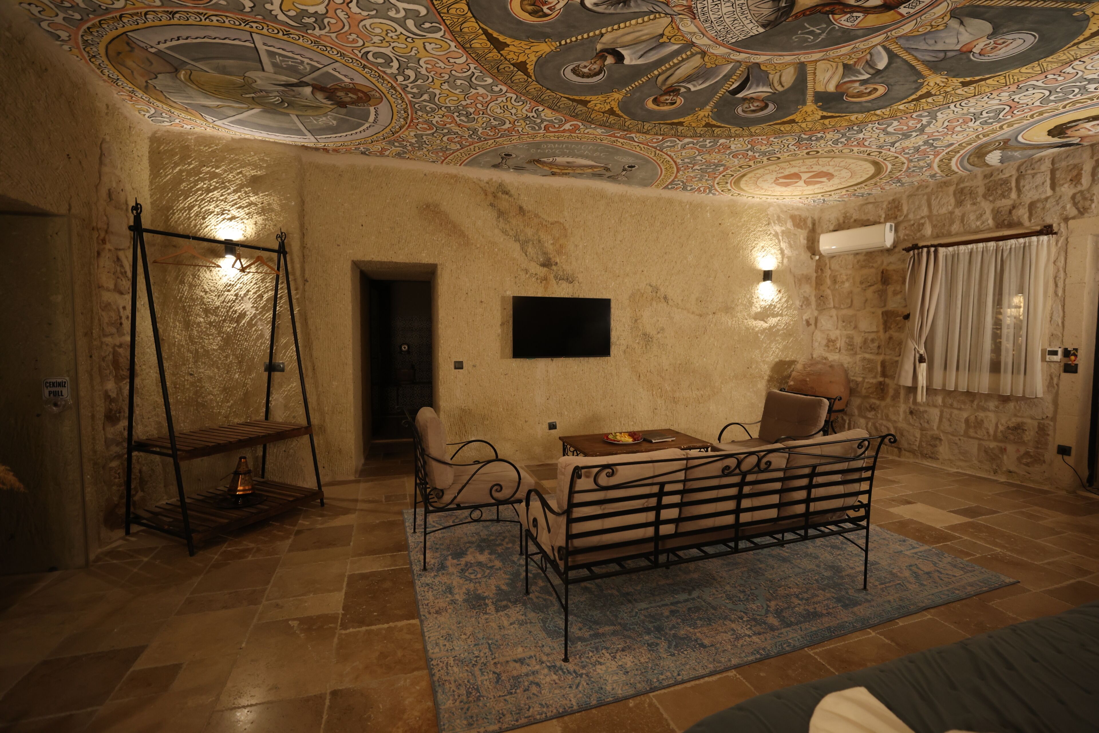 Ein Zimmer mit Steinwand, einer Bank, einem Tisch, einer kunstvoll verzierten Decke und einem kleinen Regal mit einer Lampe.